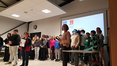 élèves de Secondes professionnels et une enseignante en charge du projet Laïcité. Les personnes sont debout dans une salle et la photo a été prise à la fin de la présentation du projet; c'est la séance des questions réponses avant la minute de silence. En fond, nous avons la dernière page d'un diaporama sur laquelle est présent le logo de la République Française et le terme Laïcité. Certains élèves sont en avant de la photo car il viennent de donner une information en chœur; d'autres arborent un sourire car ils sont heureux et soulagés du travail accomplis, le stress retombe.