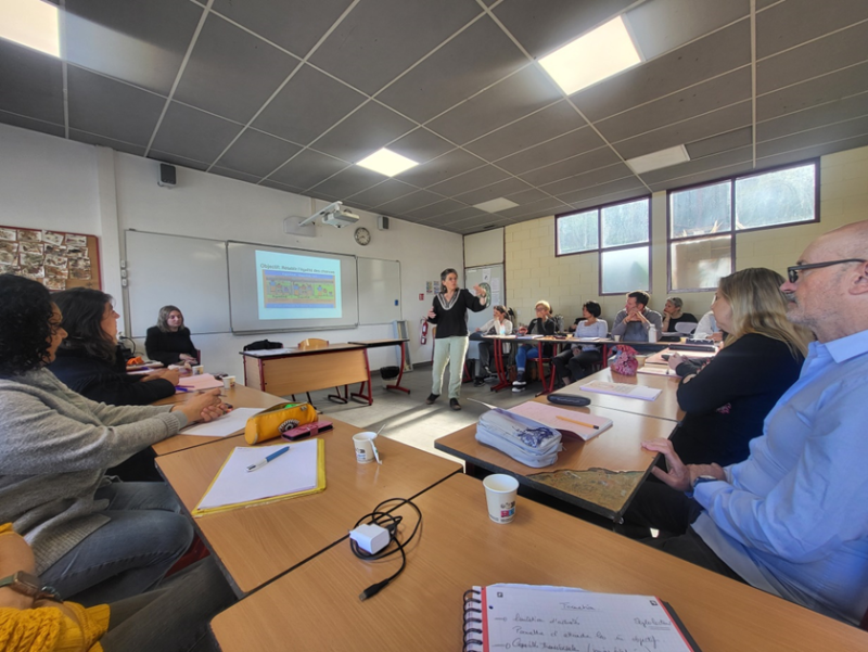 Personnels en formation. Dans une salle de classe, les personnels sont réunis autour de tables disposées en "U". En premier plan, on voit une table avec un cahier et en deuxième plan, la formatrice quii anime autour d'une présentation projettée sur un écran.